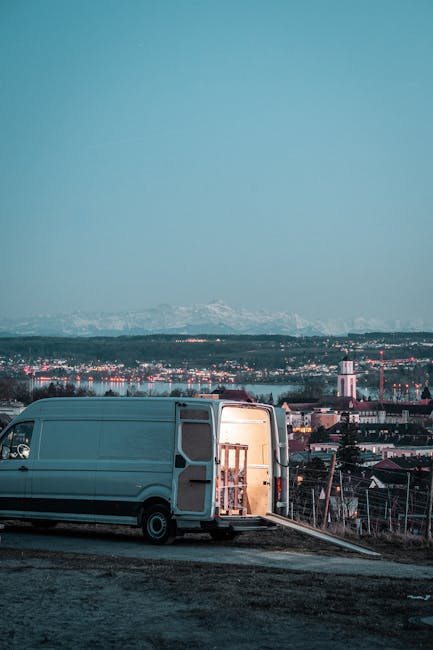 A dark red commercial pickup truck with an open cargo bed parked on an urban street. The bed is filled with various packed items for a home relocation, including cardboard boxes, fabric-covered packag
